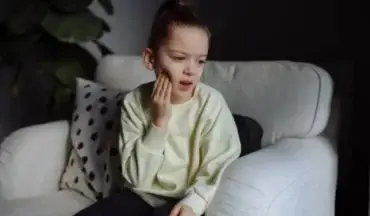 Young girl sitting on a white couch, holding her cheek in pain, possibly due to tooth pain or cavity.